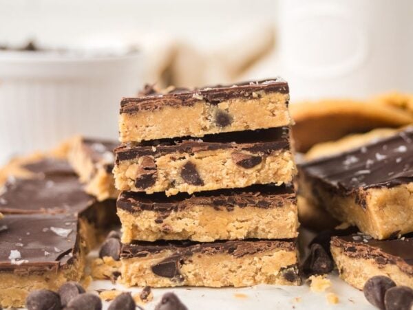 Stack of chocolate-topped Cookie Dough Bars with chocolate chips, surrounded by more bars and scattered chocolate chips.
