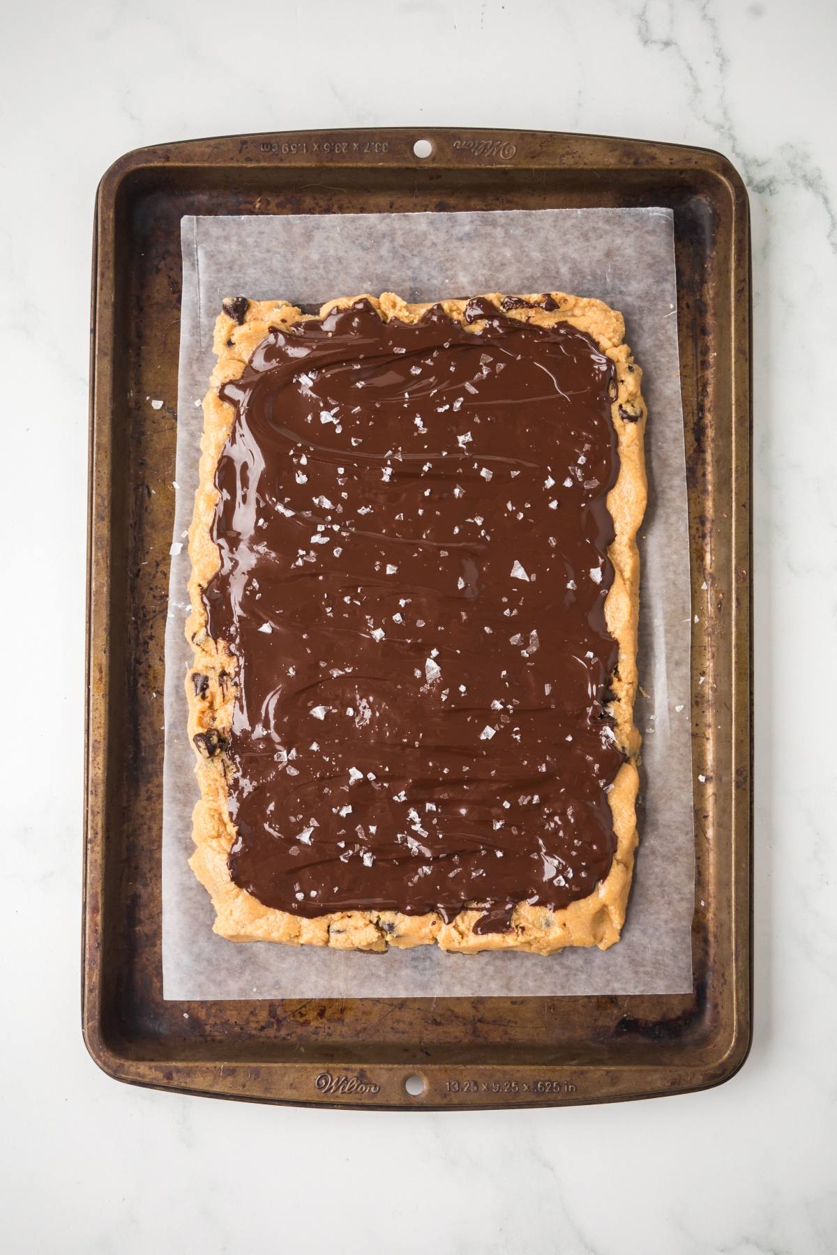Rectangular cookie topped with melted chocolate and sea salt on a baking sheet lined with parchment paper.
