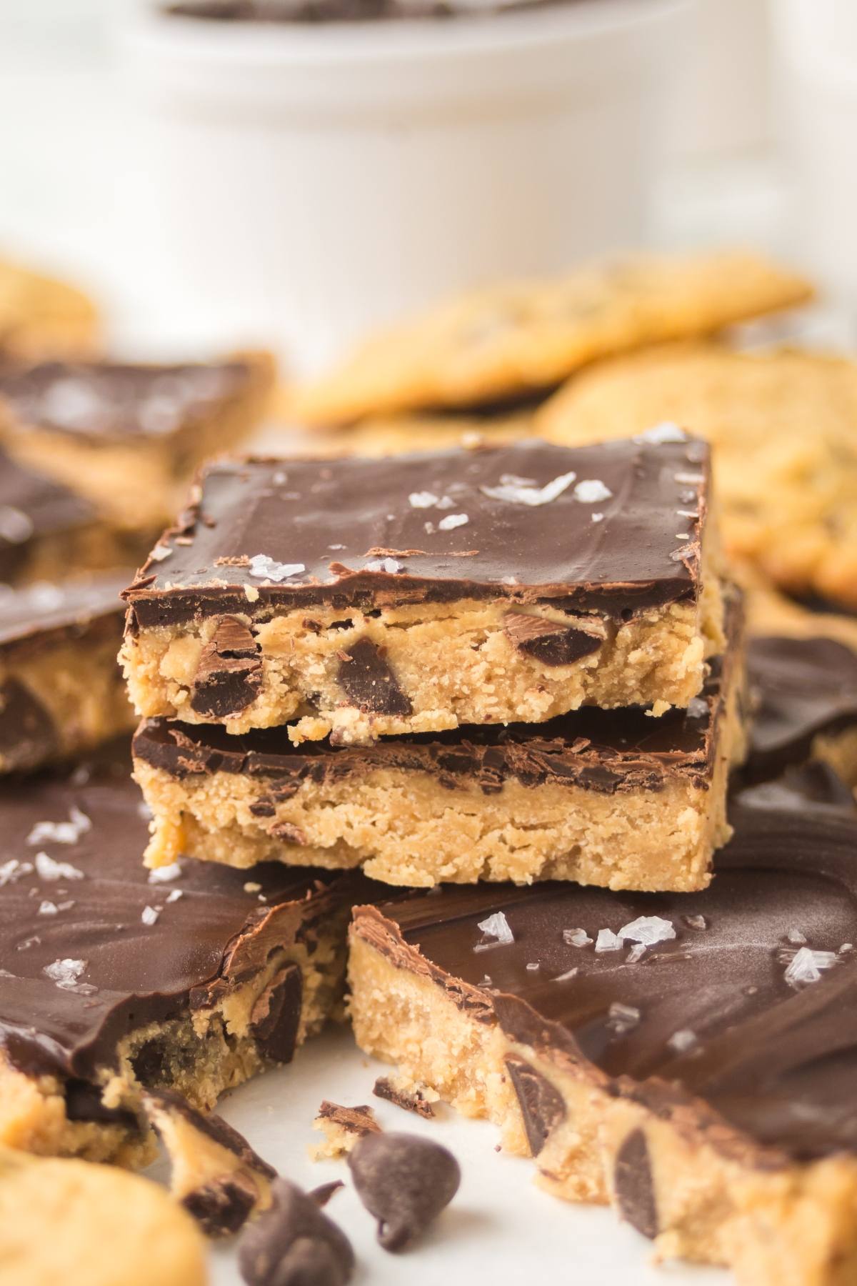 Three chocolate-topped cookie dough bars stacked, with flaky sea salt sprinkled on top and more bars in the background.