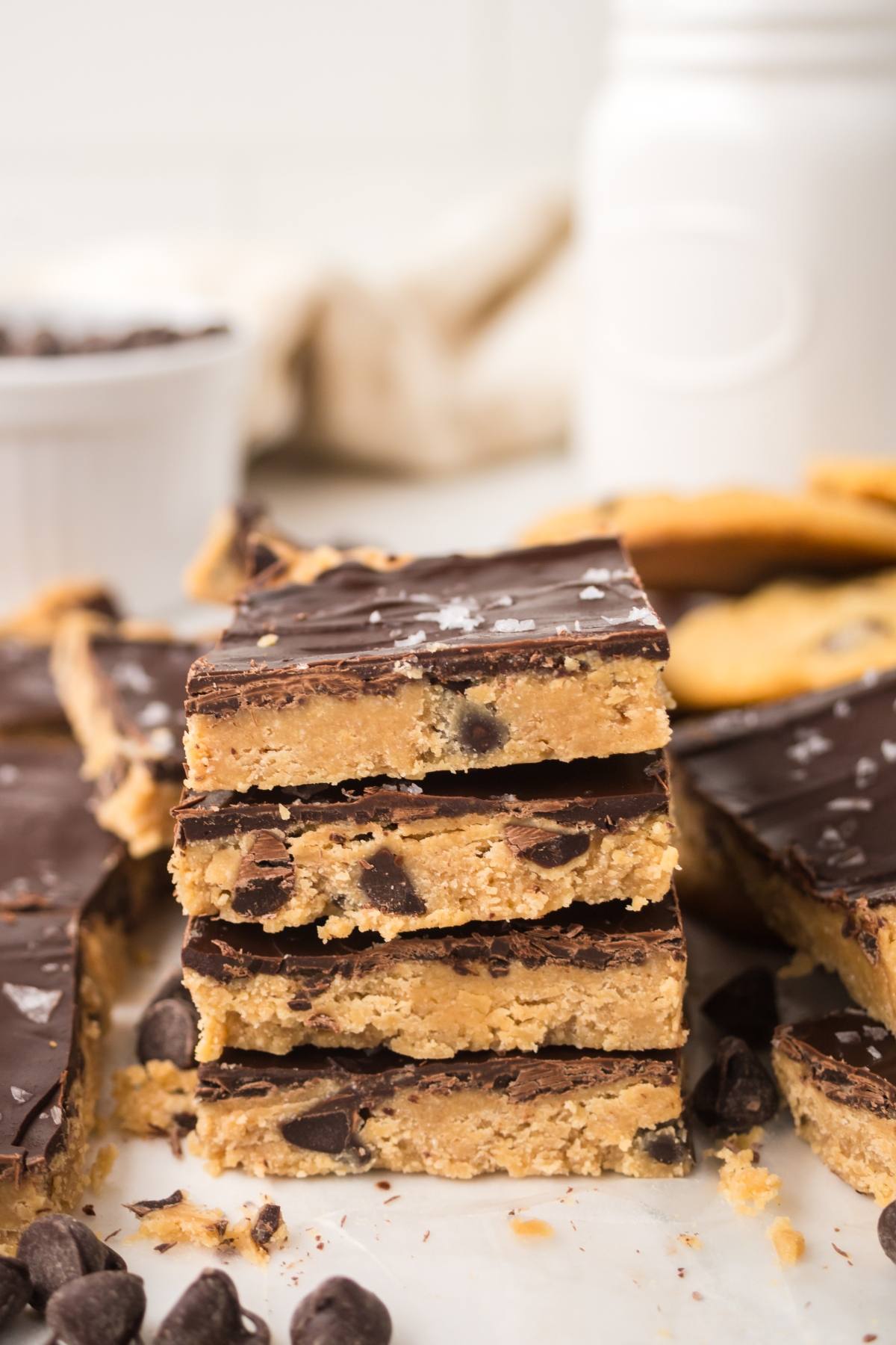 Stacked chocolate-topped cookie dough bars with chocolate chips, surrounded by more bars and scattered chips.