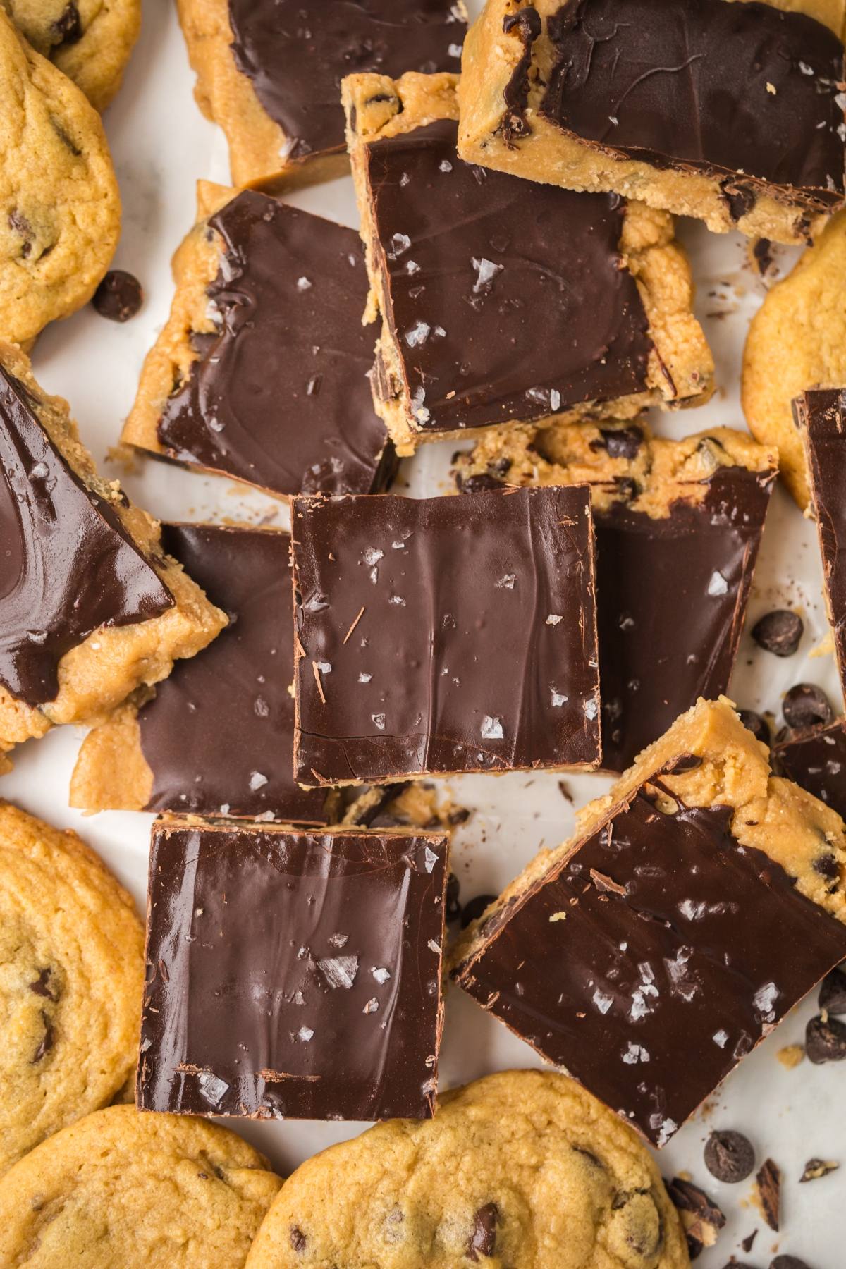 Chocolate-topped cookie dough bars and chocolate chip cookies scattered on a white surface.