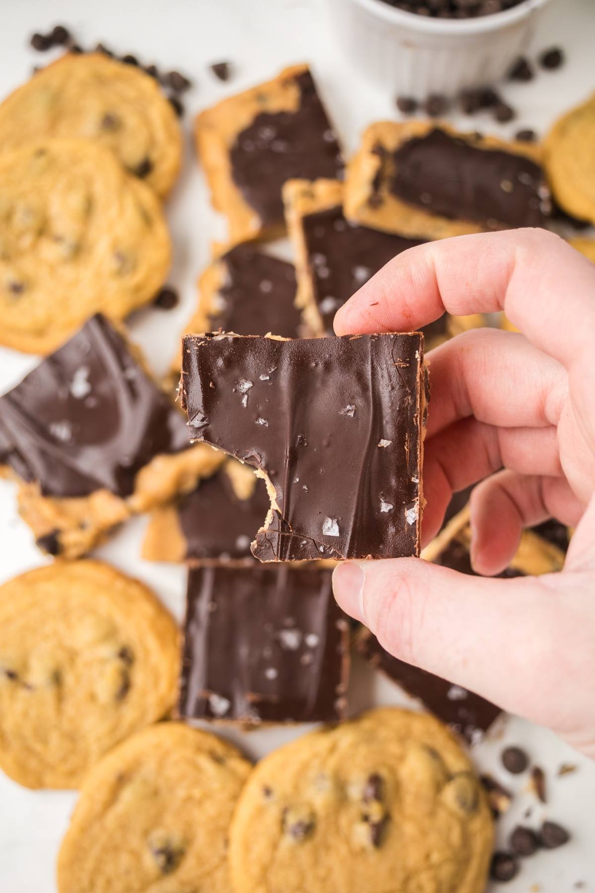 A hand holds a chocolate-covered square treat above a pile of cookies and more chocolate squares.