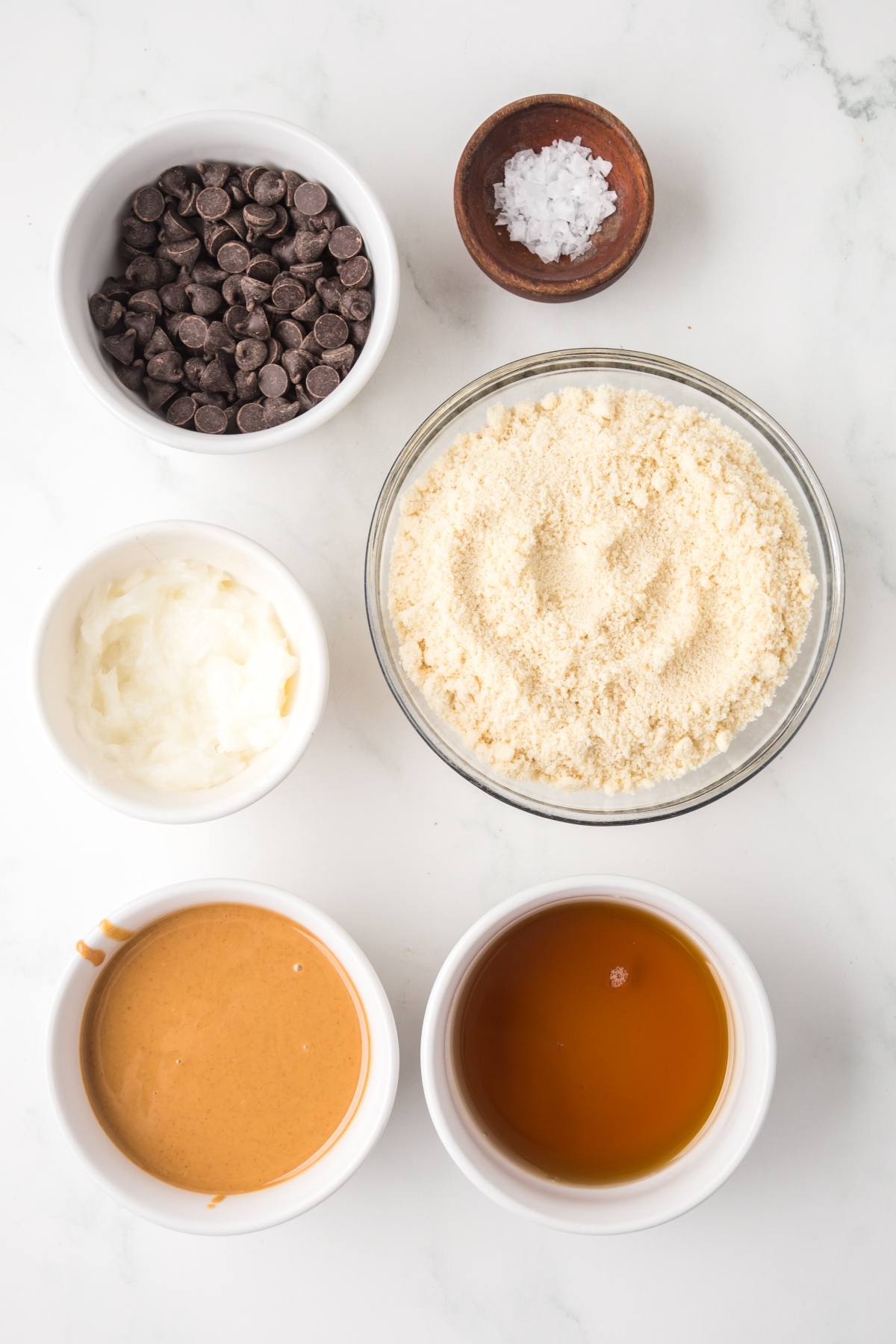 Six bowls with chocolate chips, salt, almond flour, coconut oil, peanut butter, and maple syrup on a white surface.
