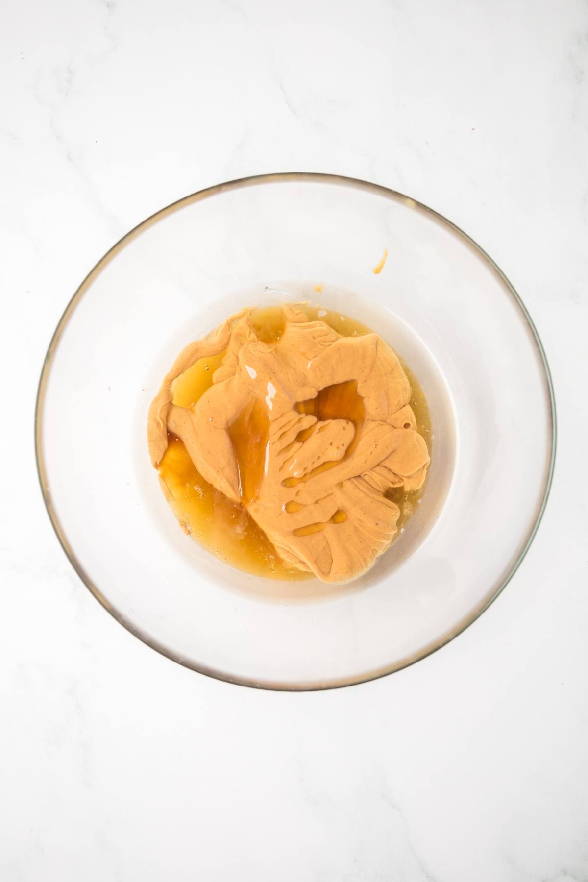 Clear glass bowl with peanut butter, honey, and vanilla on a white surface.
