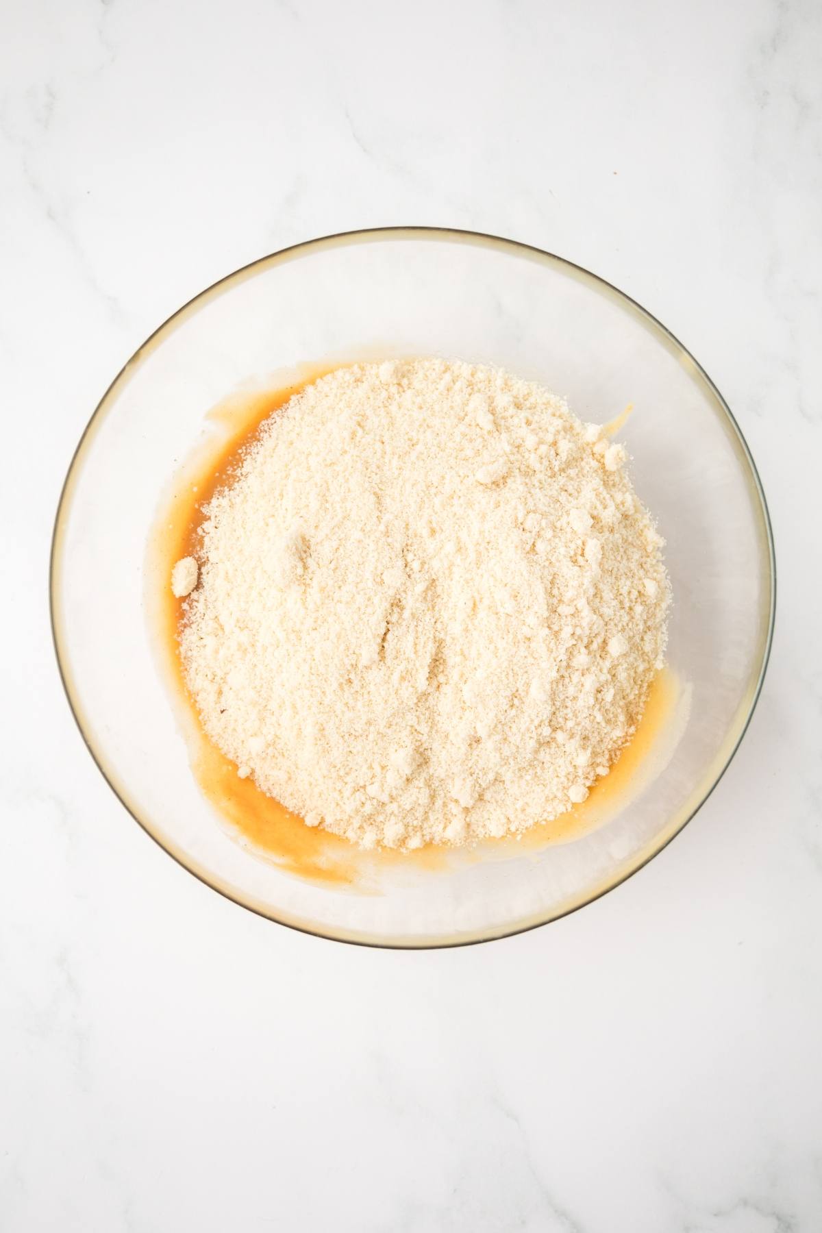 A glass bowl with almond flour on top of a yellowish liquid mixture on a white surface.
