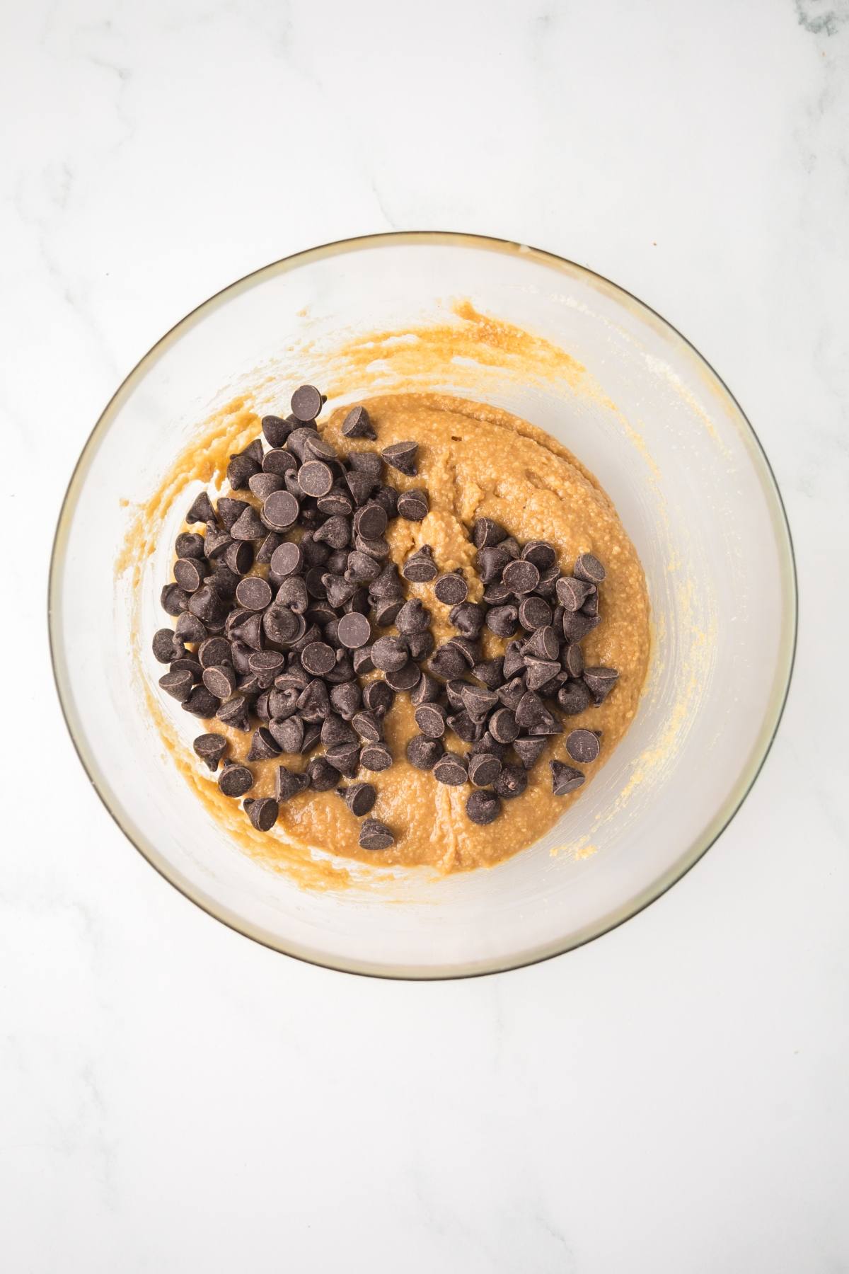 A glass bowl with cookie dough and chocolate chips on a white marble surface.