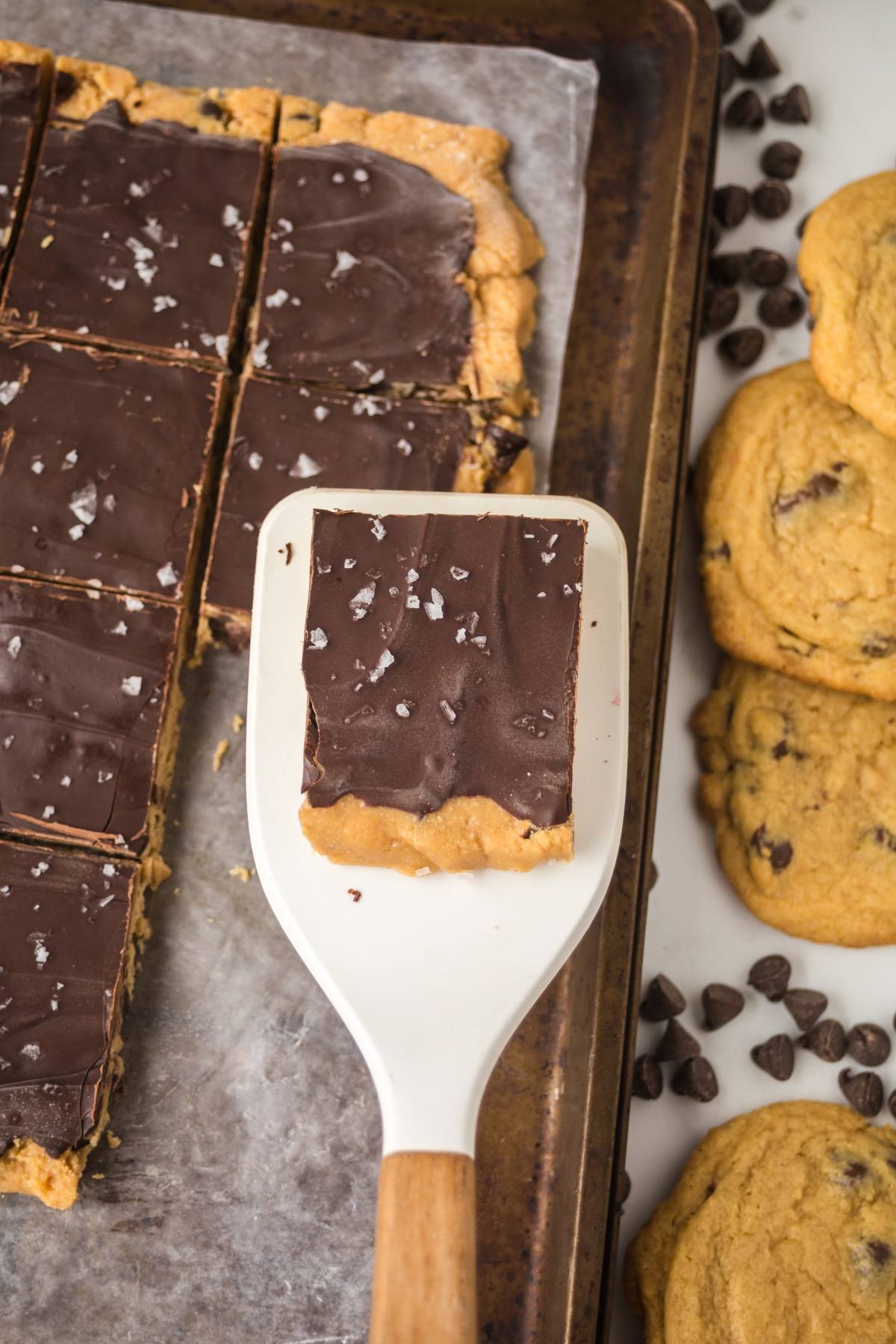 A spatula lifts a chocolate peanut butter bar topped with sea salt from a tray, next to chocolate chip cookies.