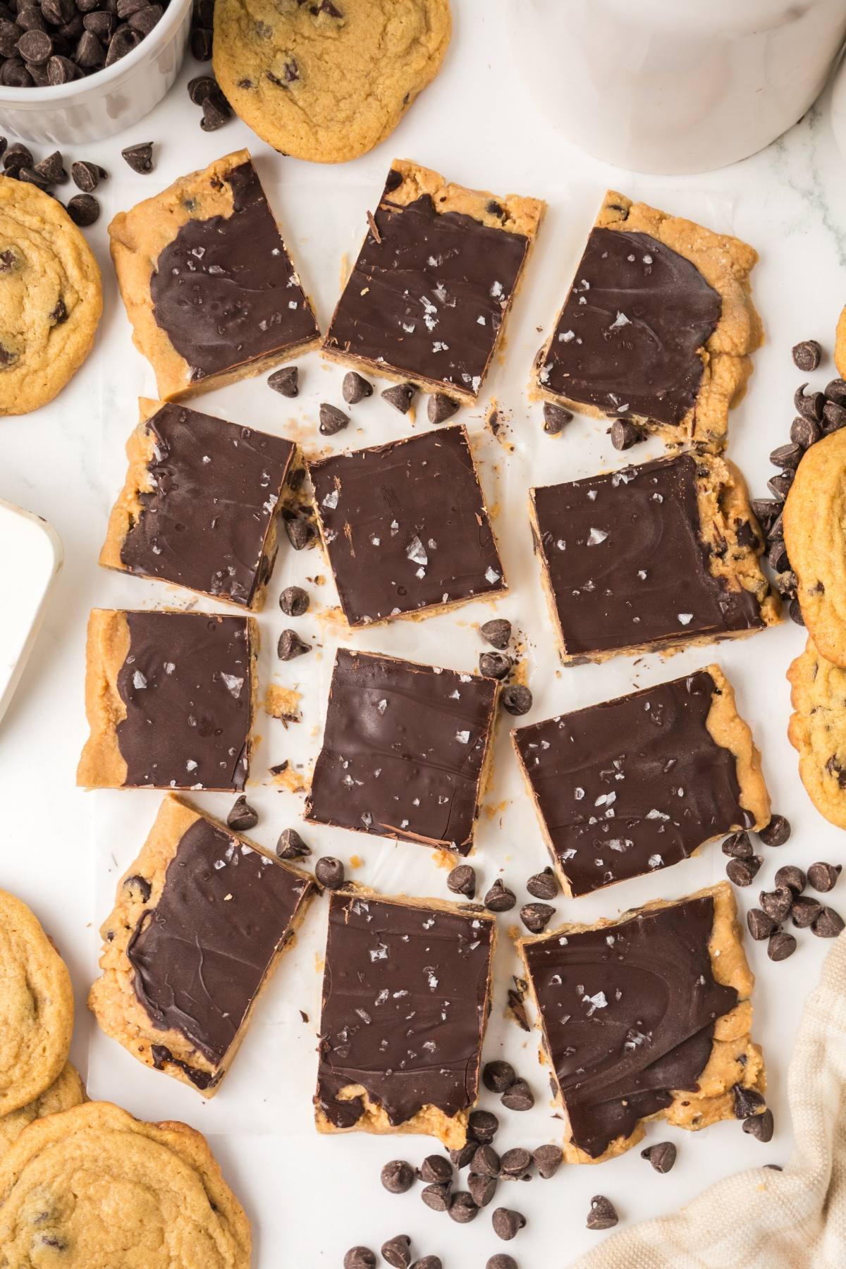 Nine chocolate-topped cookie dough bars with sea salt, surrounded by cookies and chocolate chips on a white surface.
