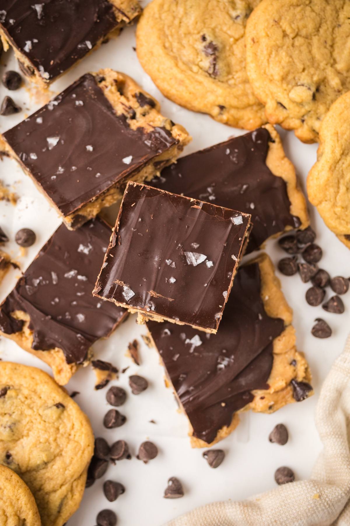 Chocolate chip cookies and chocolate-topped cookie dough bars are surrounded by scattered chocolate chips and sea salt.