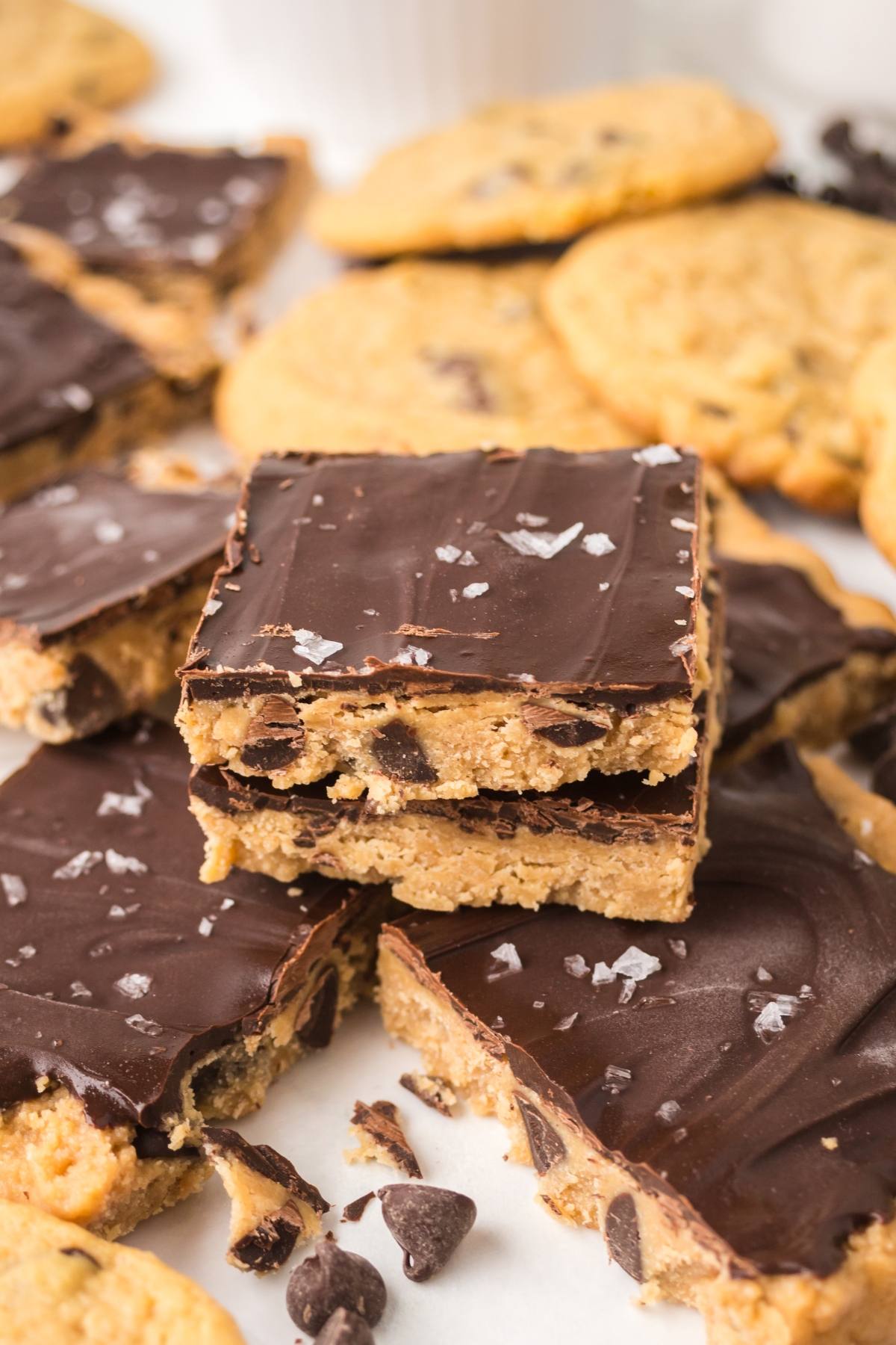 Chocolate-topped cookie dough bars with chocolate chips, stacked with chocolate chip cookies in the background.