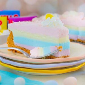 A pastel rainbow layered cheesecake slice with whipped cream, inspired by a Layered Jello Pie, with Jello boxes and candies in the background.