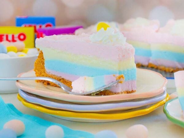 A pastel rainbow layered cheesecake slice with whipped cream, inspired by a Layered Jello Pie, with Jello boxes and candies in the background.