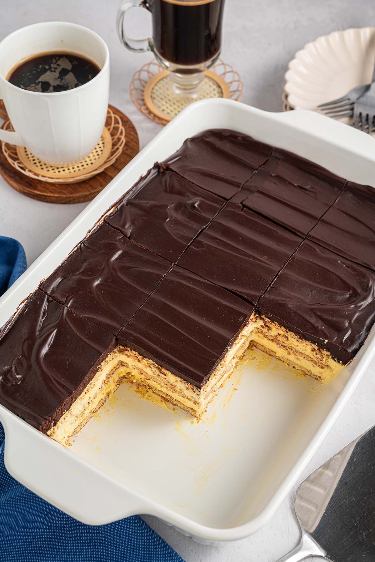 Rectangular Eclair Cake layered dessert with chocolate topping, partly sliced, sits next to cups of coffee on a table.