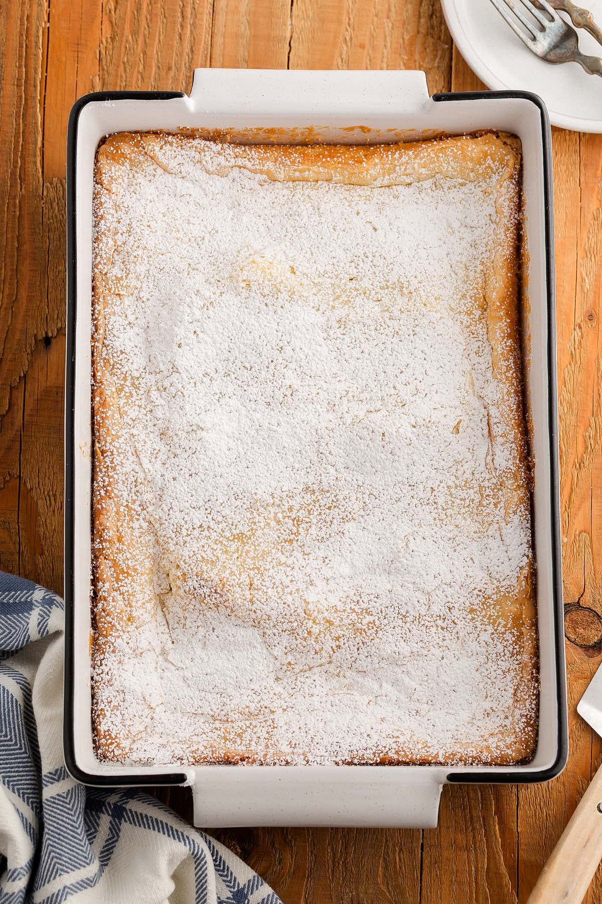 Golden gooey butter cake in a white baking dish, topped with a generous layer of powdered sugar on a wooden table.