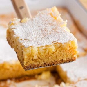 A spatula lifts a square piece of gooey butter cake, dusted with powdered sugar, from a baking dish, revealing its golden, creamy layers.