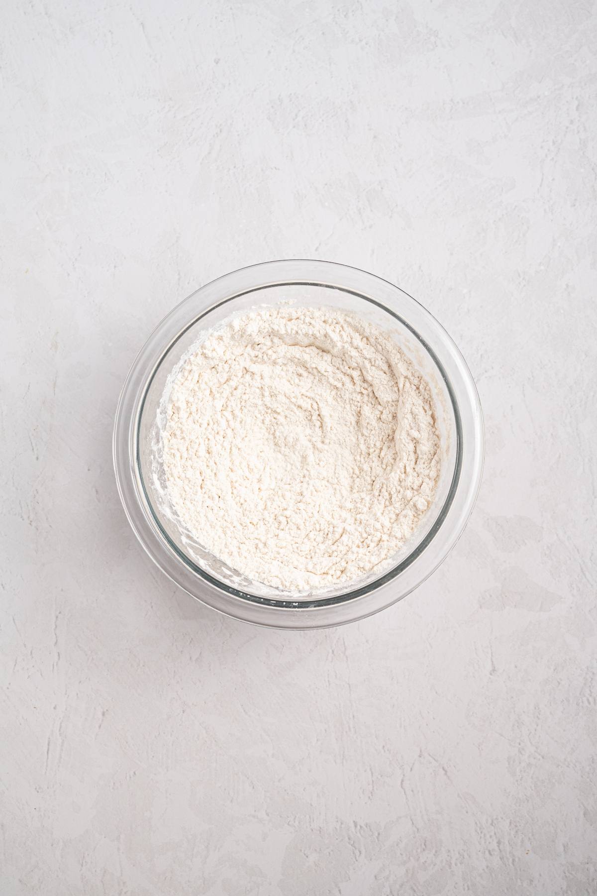 A clear glass bowl filled with white flour on a light textured surface.