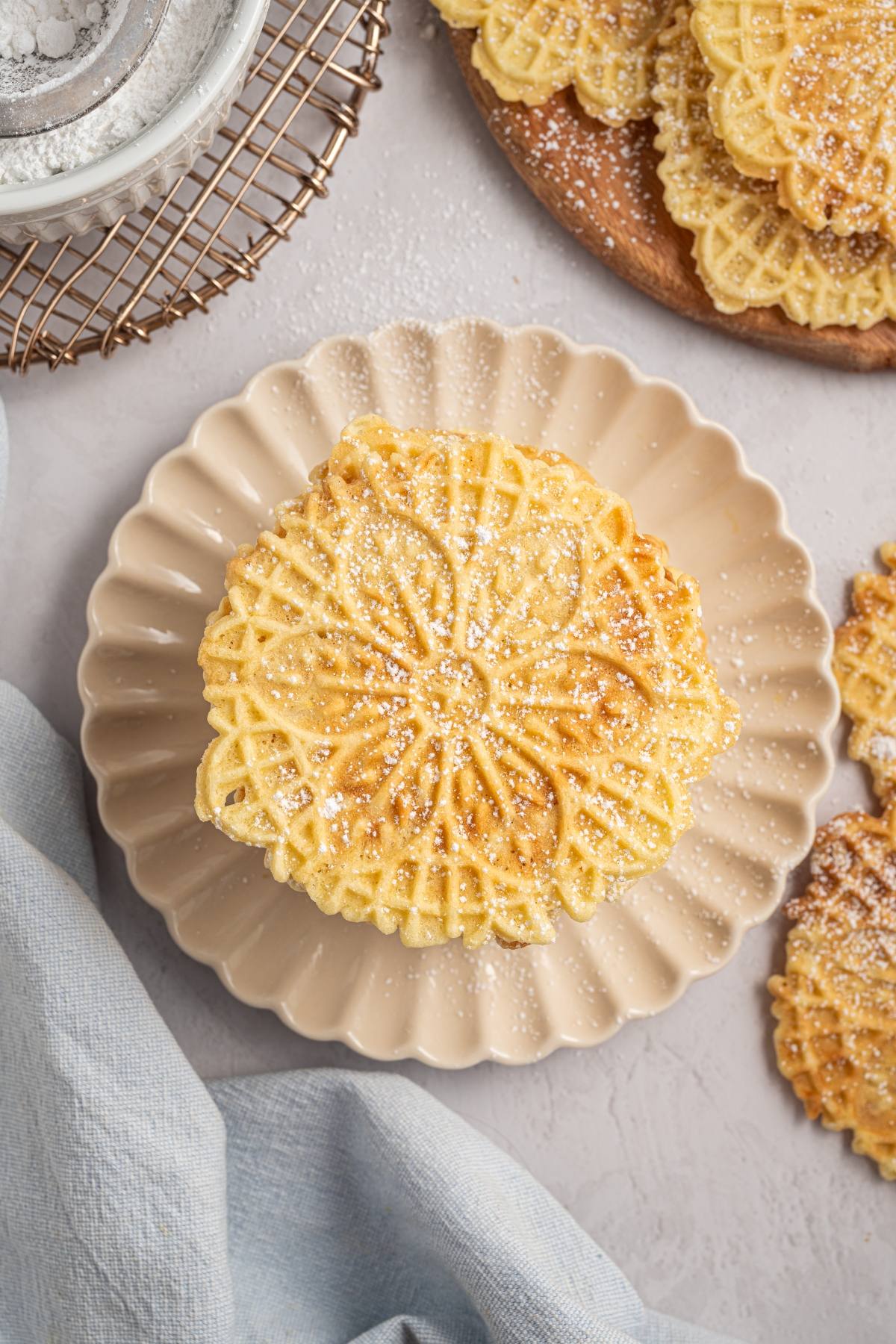 A plate with a stack of powdered sugar-dusted italian pizzelle cookies on a light, textured surface.