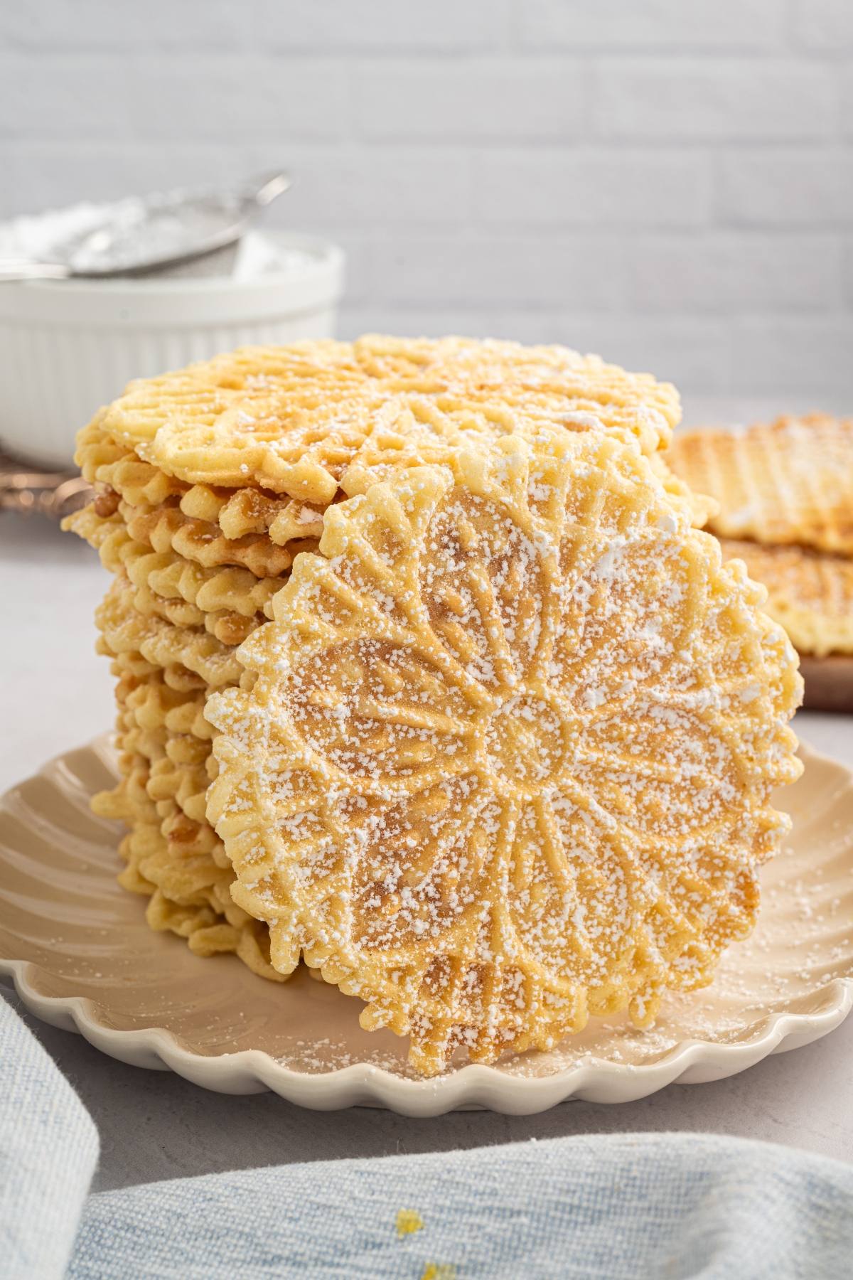 A stack of round, patterned italian pizzelle cookies dusted with powdered sugar on a beige plate.