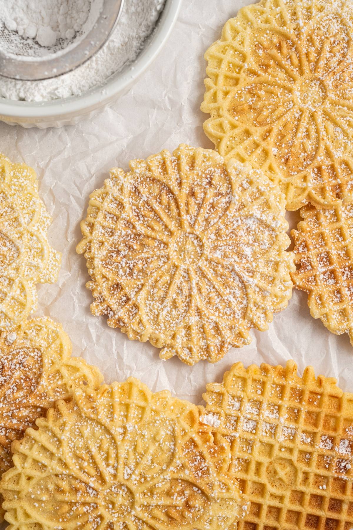 Round, patterned italian pizzelle cookies dusted with powdered sugar on crinkled parchment paper.