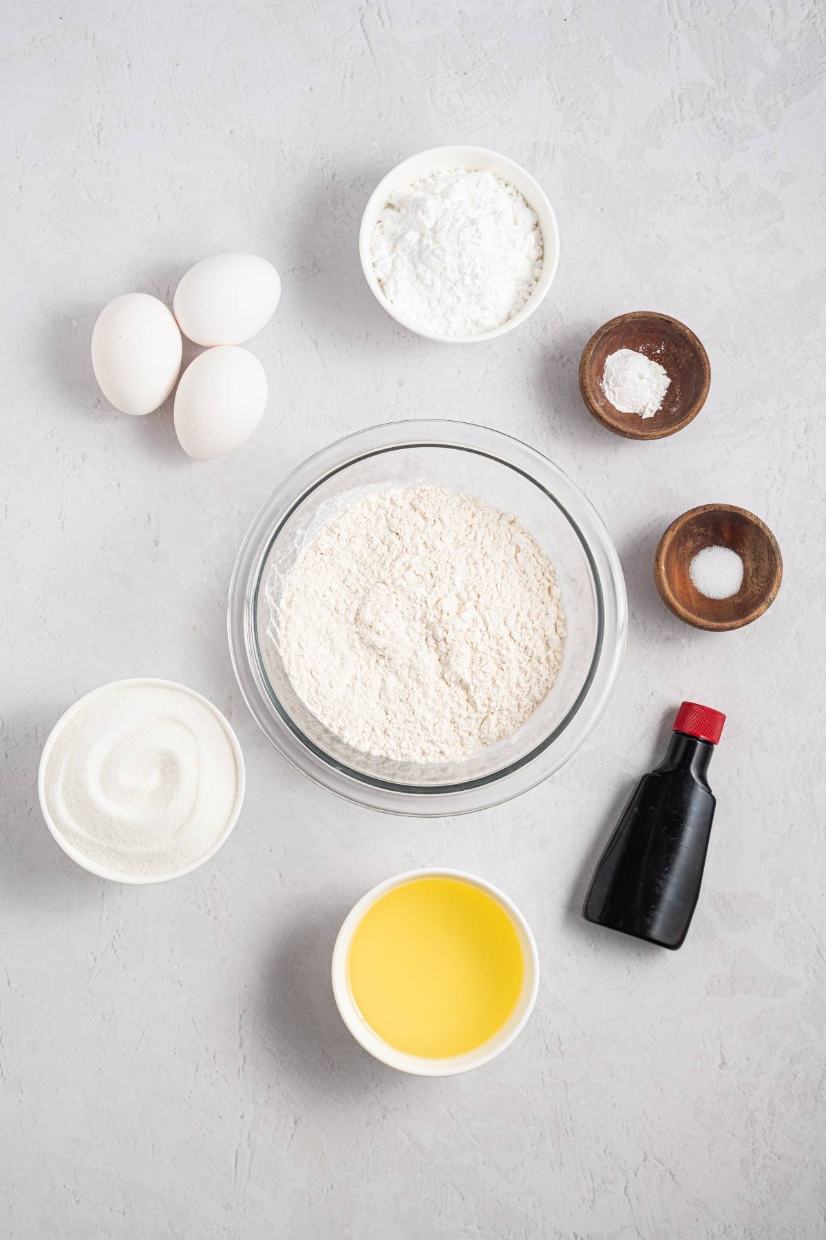 Baking ingredients: flour, eggs, sugar, oil, vanilla, salt, baking powder, and powdered sugar on a gray background.