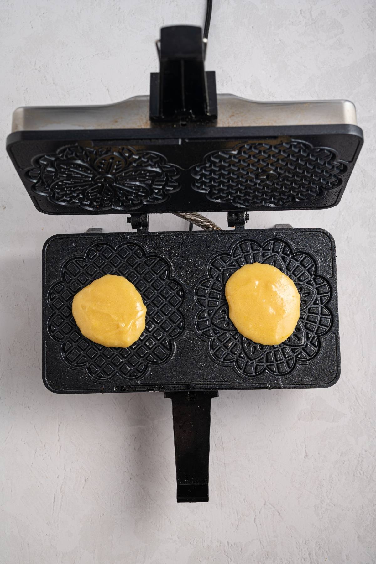 Open pizzelle maker with two portions of yellow dough placed on decorative black plates, ready to cook.