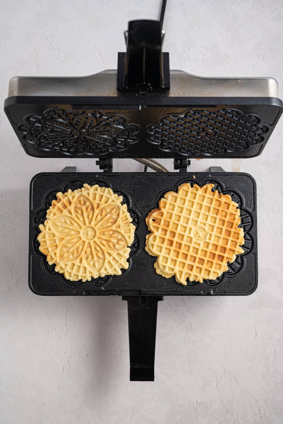 An open pizzelle maker with two golden pizzelle cookies inside, viewed from above.