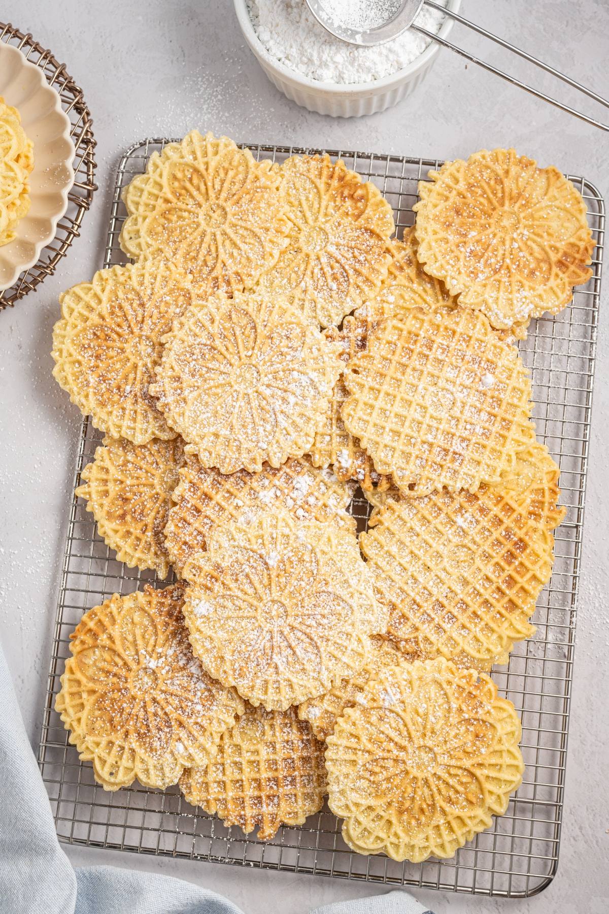 A tray of round, patterned italian pizzelle cookies dusted with powdered sugar on a wire rack.