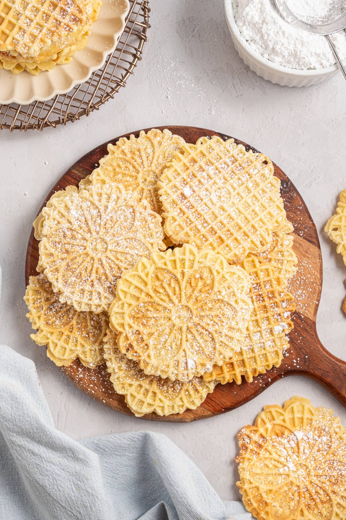 Round Italian pizzelle cookies dusted with powdered sugar on a wooden board and plate.