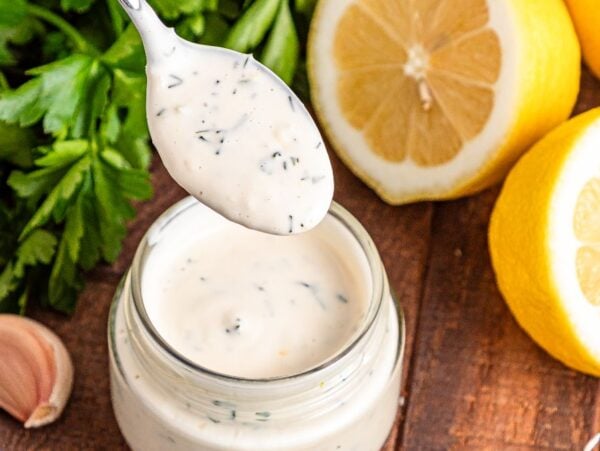 A spoonful of creamy Lemon Aioli dressing above a jar, with fresh lemons, parsley, and garlic on a wooden table.