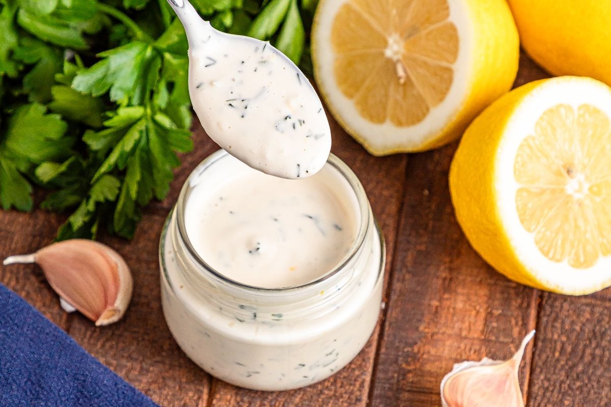 A jar of creamy herb Lemon Aioli with a spoon, fresh parsley, garlic, and halved lemons on a wooden surface.