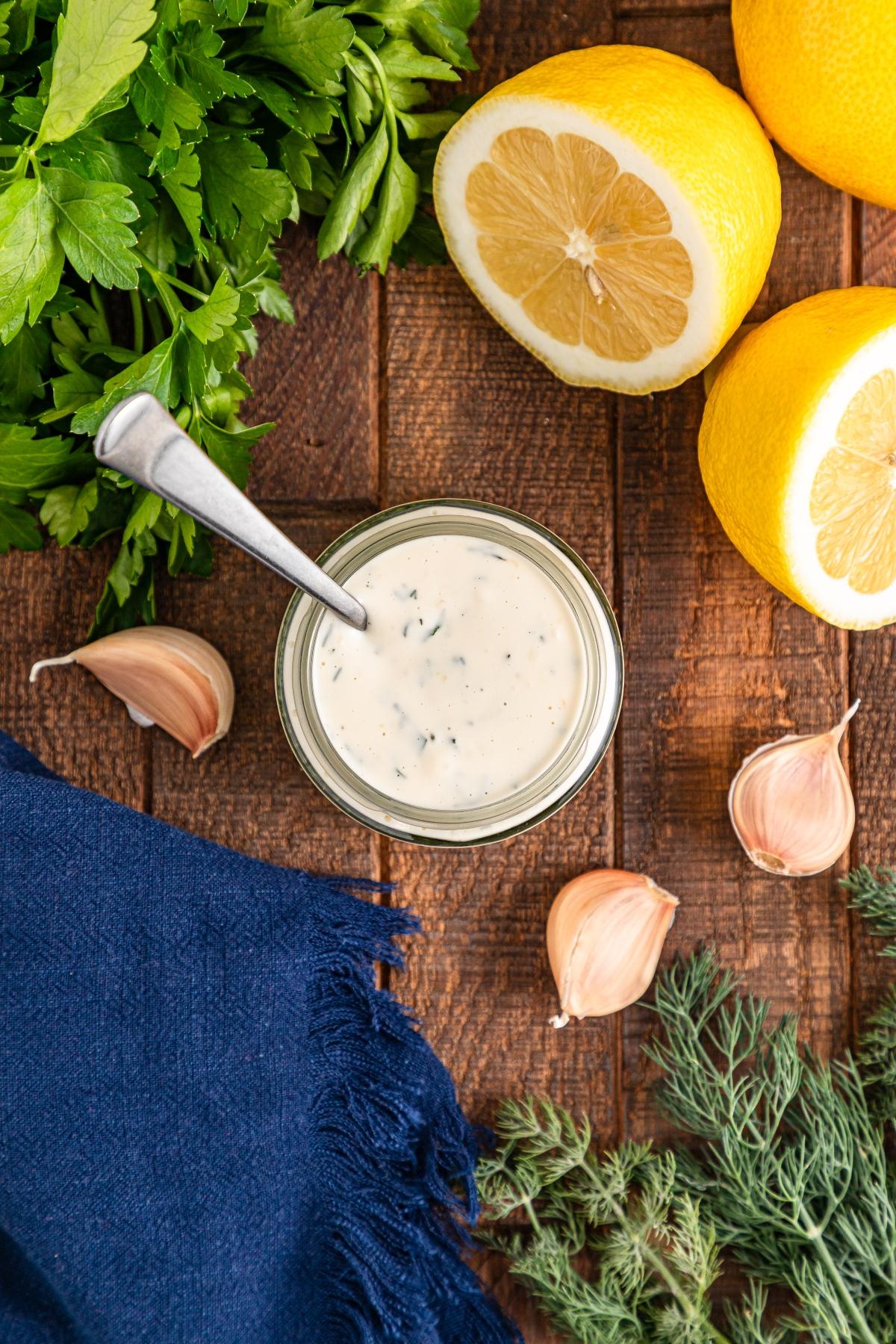 Jar of creamy ranch dressing with a spoon, surrounded by lemon aioli halves, garlic, fresh herbs, and a blue napkin.