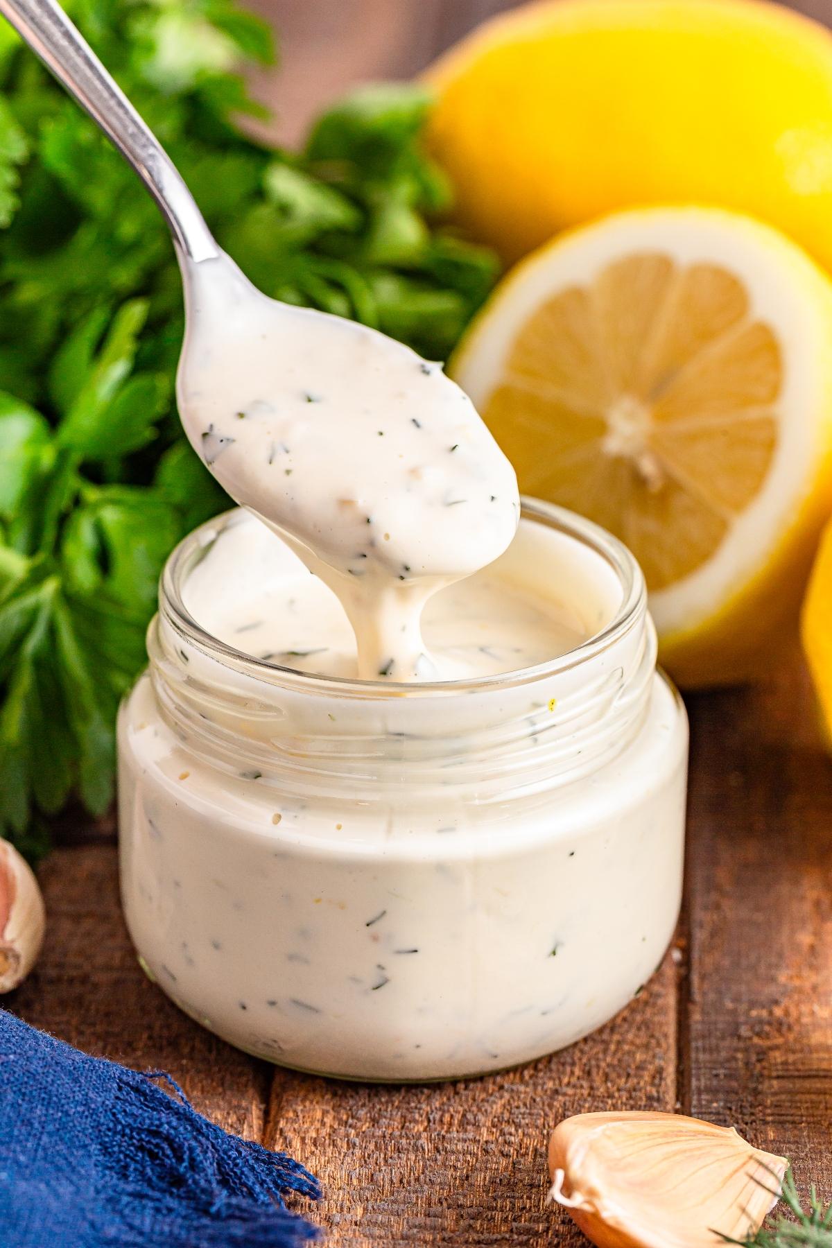 A spoon scoops creamy herb dressing from a jar, with lemons, parsley, and garlic in the background.