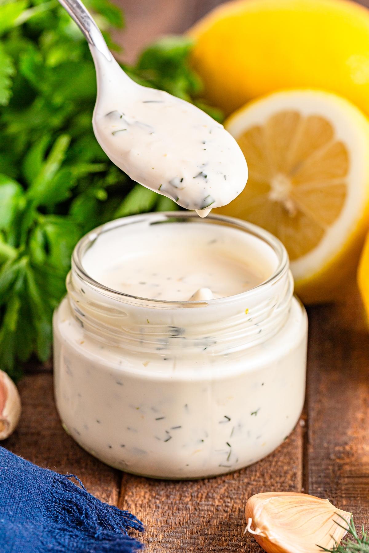 A spoon lifts creamy herb sauce from a jar, with lemons, parsley, and garlic in the background.