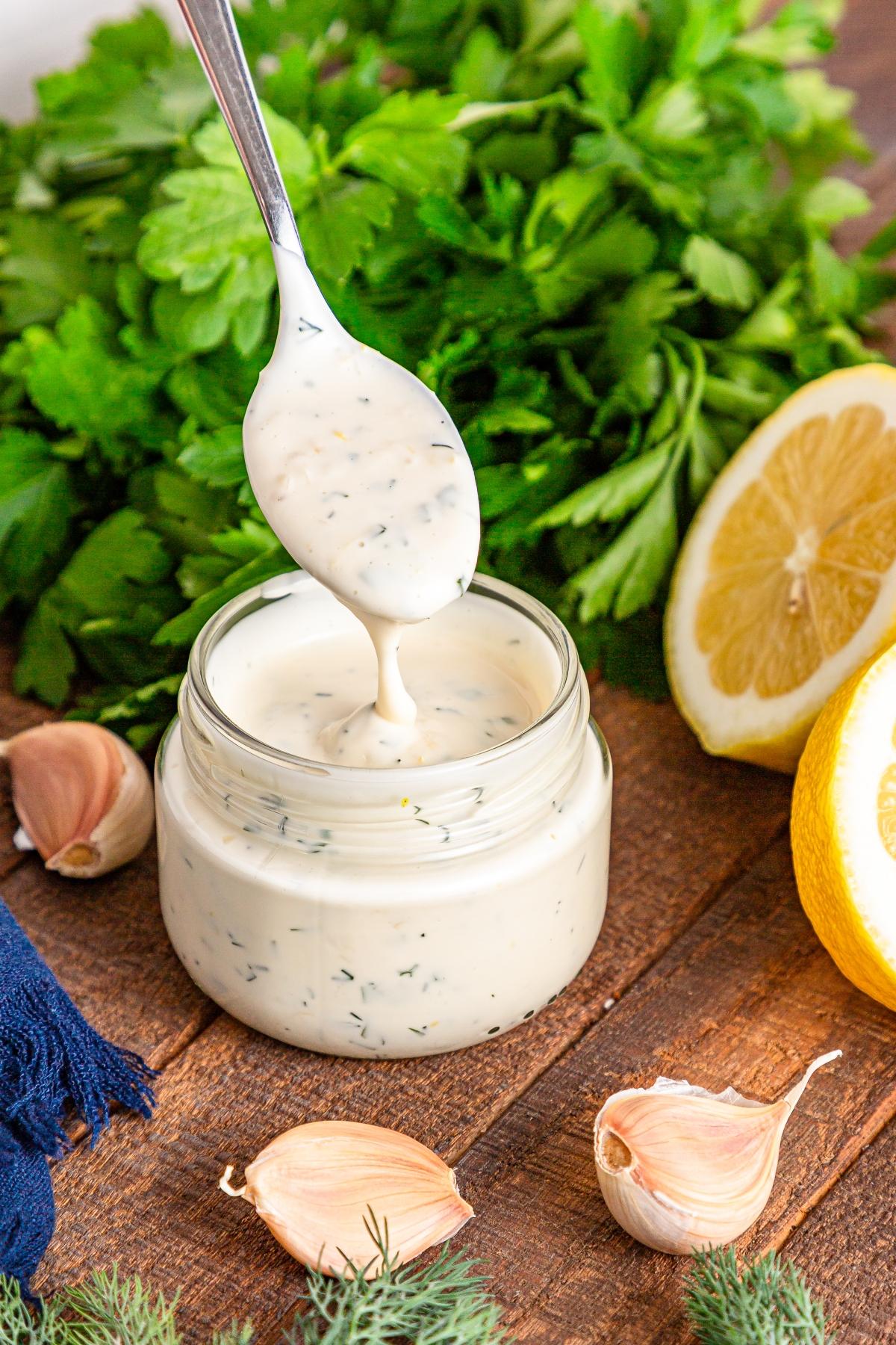 A spoon lifts creamy herb dressing from a jar, surrounded by garlic, lemon, and fresh parsley.
