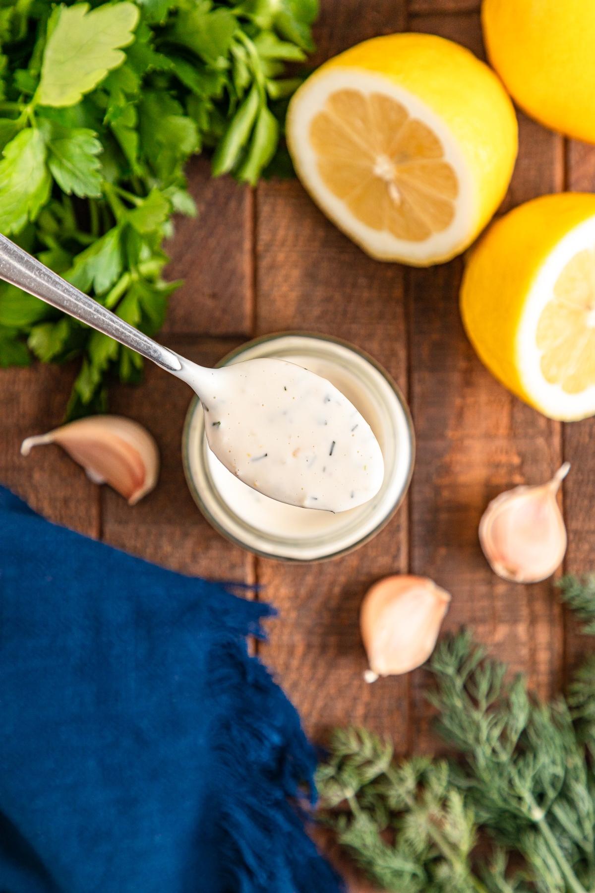 Spoonful of creamy ranch dressing over a jar, surrounded by lemons, garlic, parsley, and dill on a wooden table.