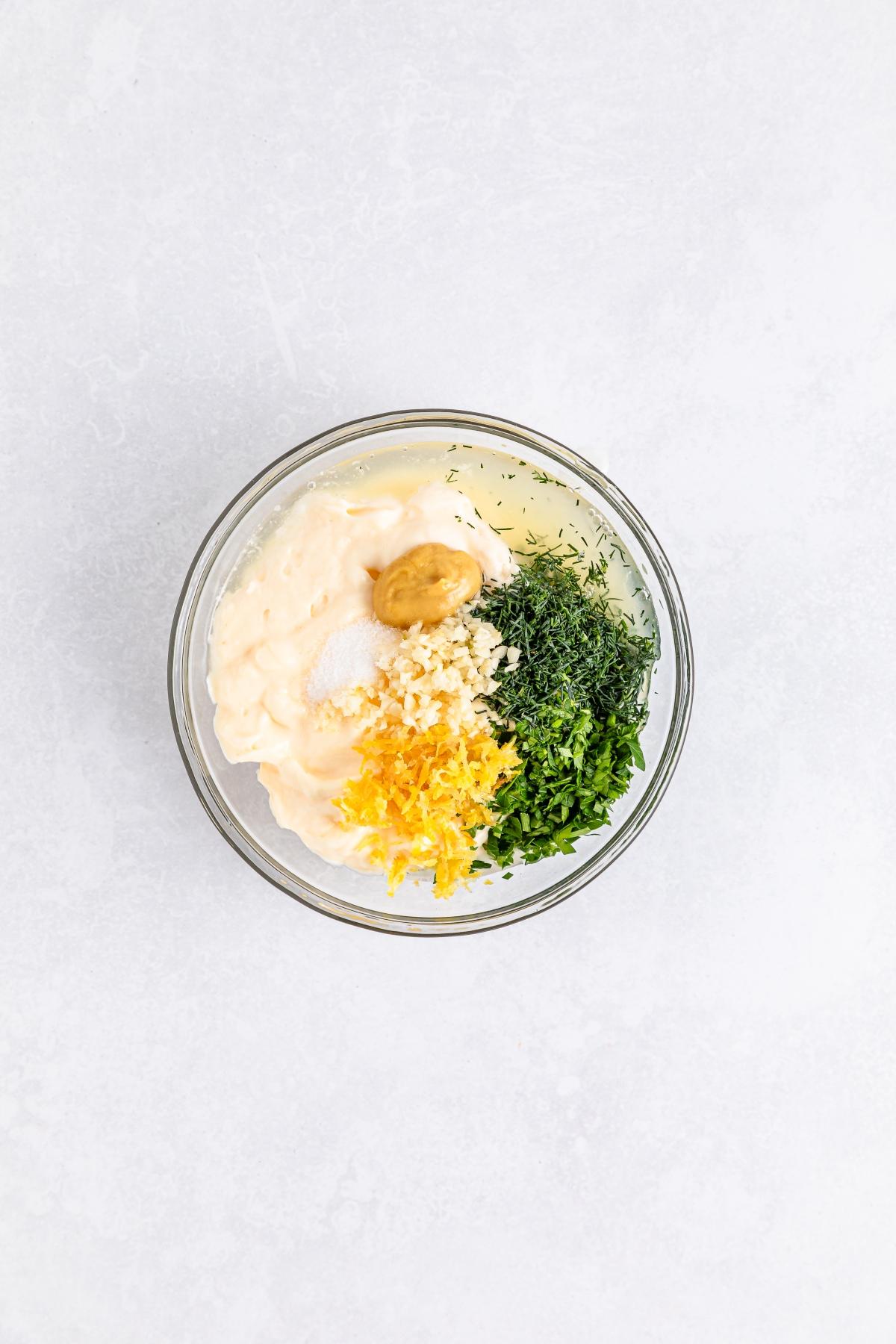Glass bowl with mayonnaise, chopped herbs, minced garlic, mustard, lemon zest, and salt on a light surface.