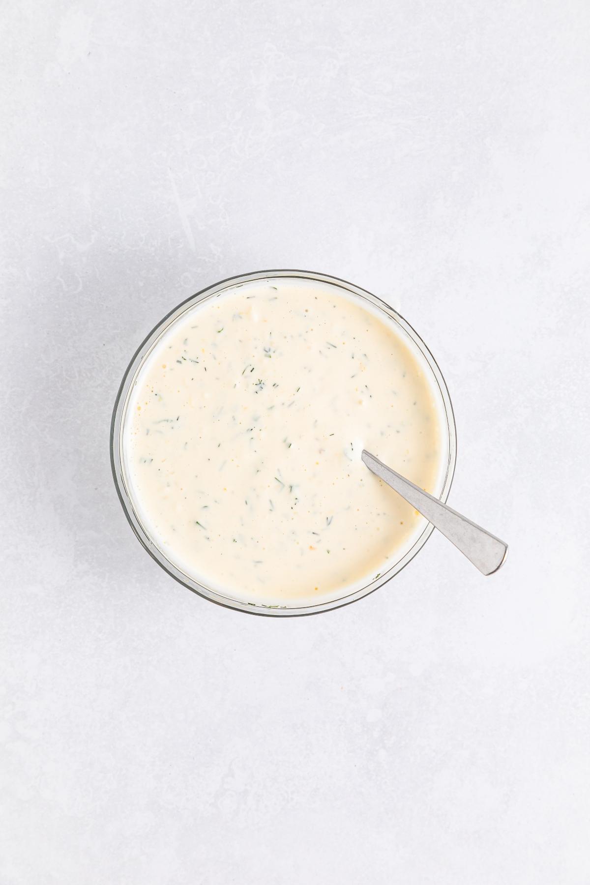 A bowl of creamy ranch dressing with herbs, with a spoon resting inside, on a light background.