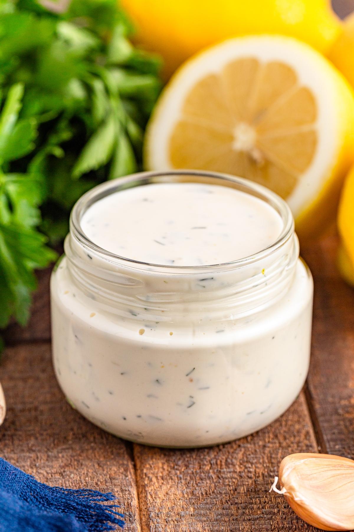 A jar of creamy ranch dressing with herbs, surrounded by parsley, lemon, and a clove of garlic.