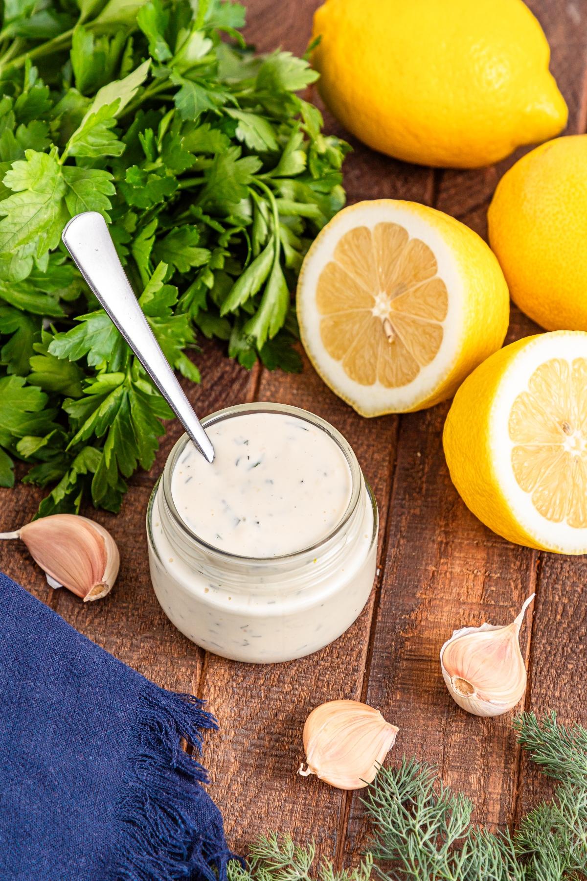 Jar of creamy herb sauce with a spoon, surrounded by parsley, lemons, garlic cloves, and dill on a wooden table.