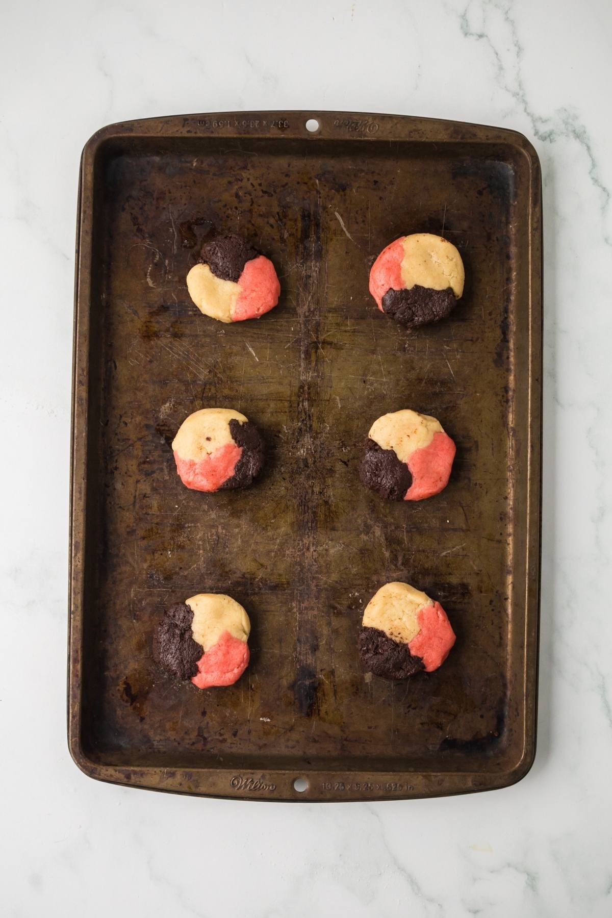 Six tri-color cookies with chocolate, vanilla, and pink dough on a worn baking sheet, on a white surface.