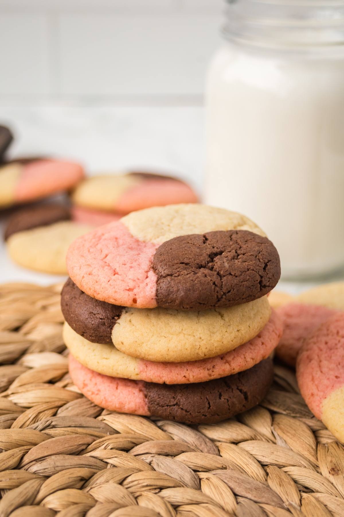 Three-tone neapolitan  cookies stacked on a woven mat, with a jar of milk and more cookies in the background.