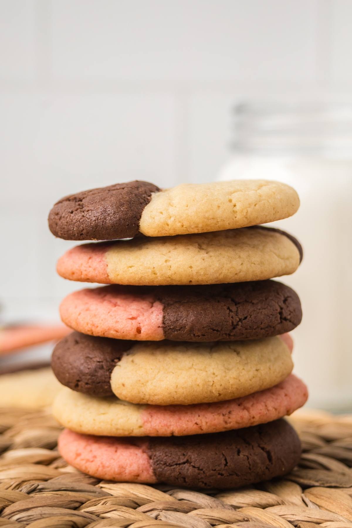 A stack of chocolate, vanilla, and strawberry neapolitan  cookies next to a glass of milk on a woven mat.