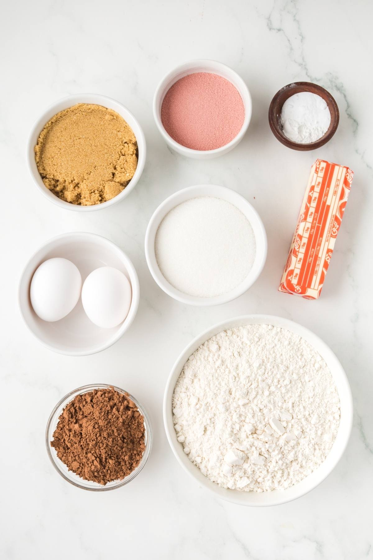 Baking ingredients: eggs, flour, cocoa, sugars, butter, baking soda, and pink powder in bowls on a white surface.
