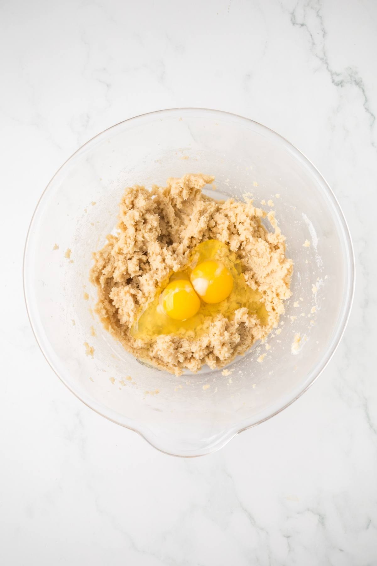 A glass bowl with cookie dough mixture and two raw eggs on top, on a white marble surface.