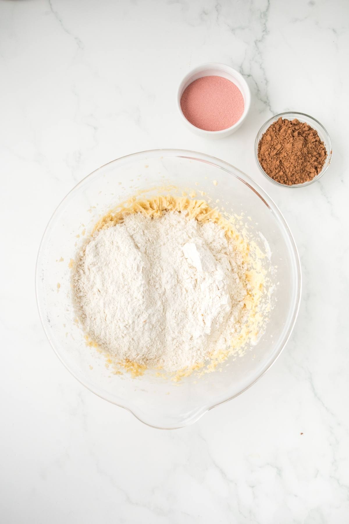 A glass bowl with flour mixture, cocoa powder, and pink sugar in small bowls on a white surface.