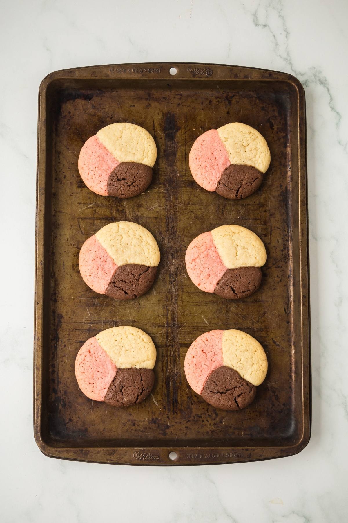 Six tri-colored neapolitan cookies&mdash;pink, yellow, and brown&mdash;on a dark baking sheet, viewed from above.