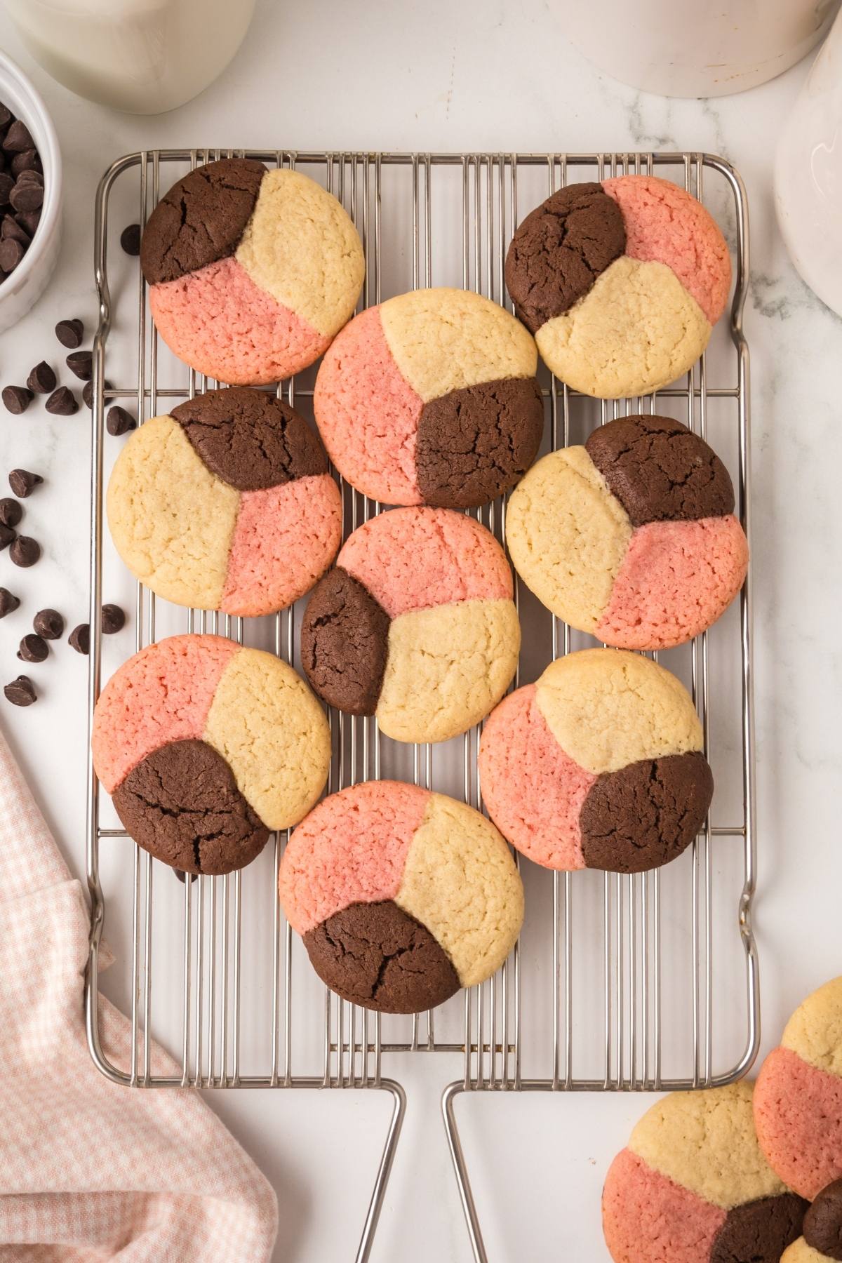 Round neapolitan  cookies with pink, brown, and beige sections cooling on a wire rack, chocolate chips nearby.