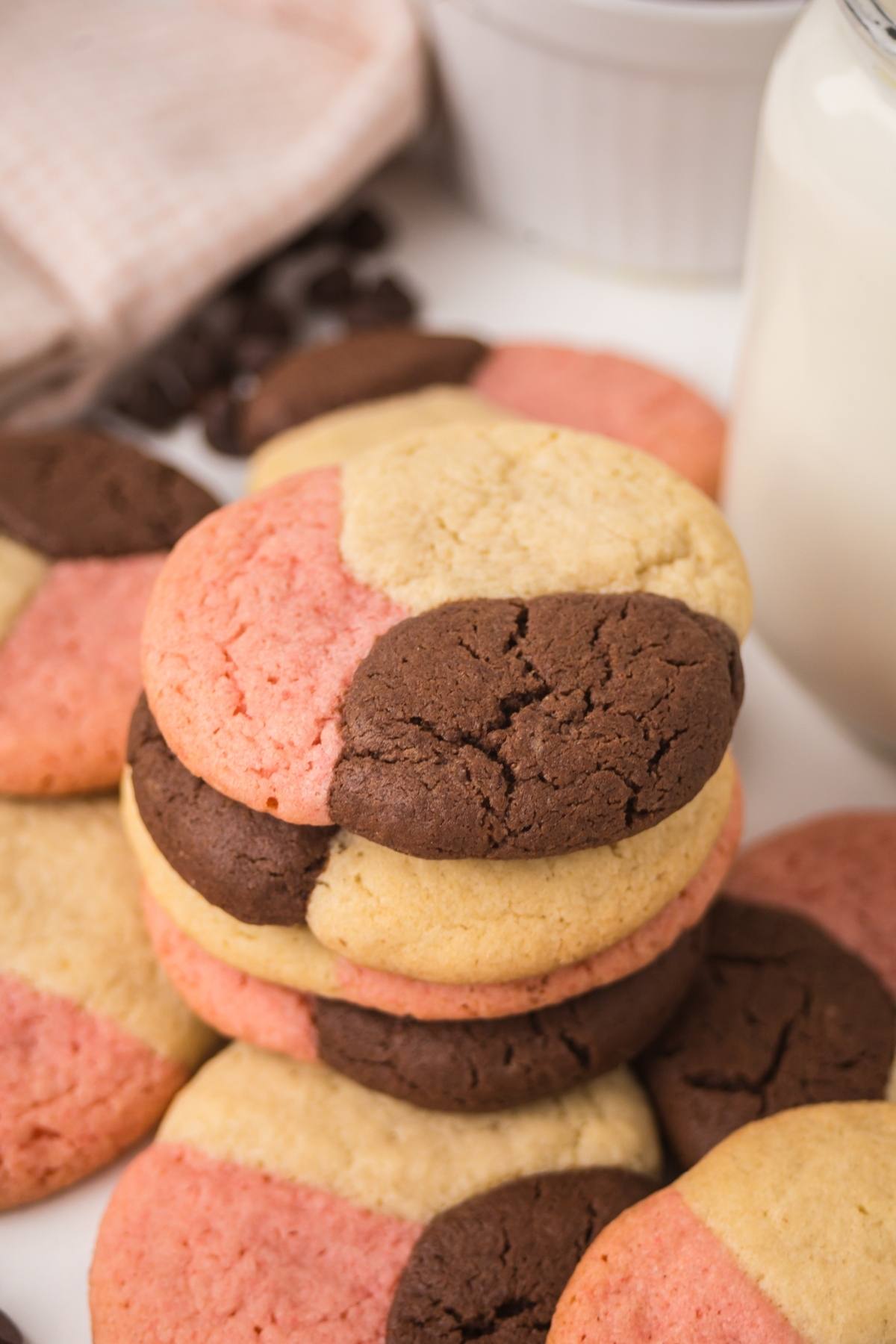 A stack of tri-color neapolitan  cookies in pink, chocolate, and vanilla next to a glass of milk.