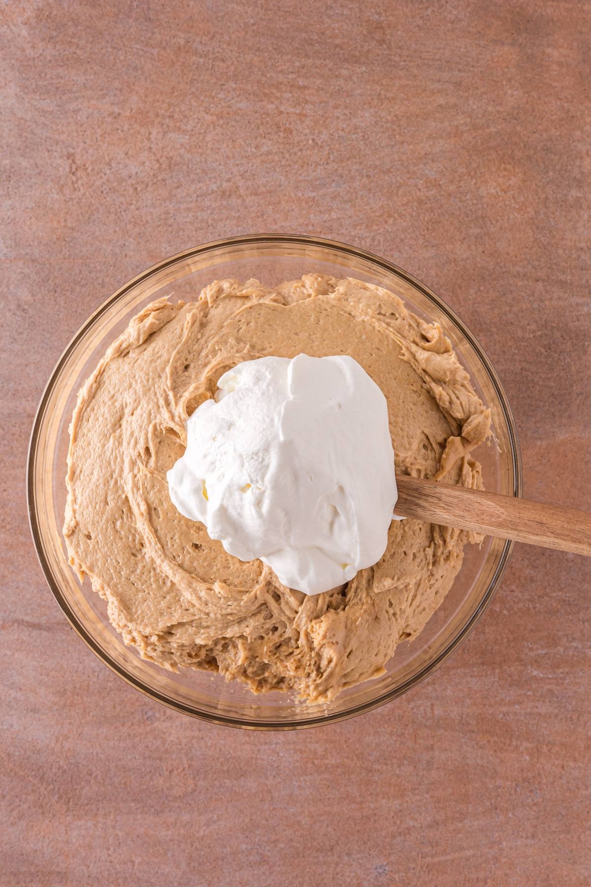 A glass bowl with brown batter and a dollop of whipped cream being mixed with a wooden spatula.