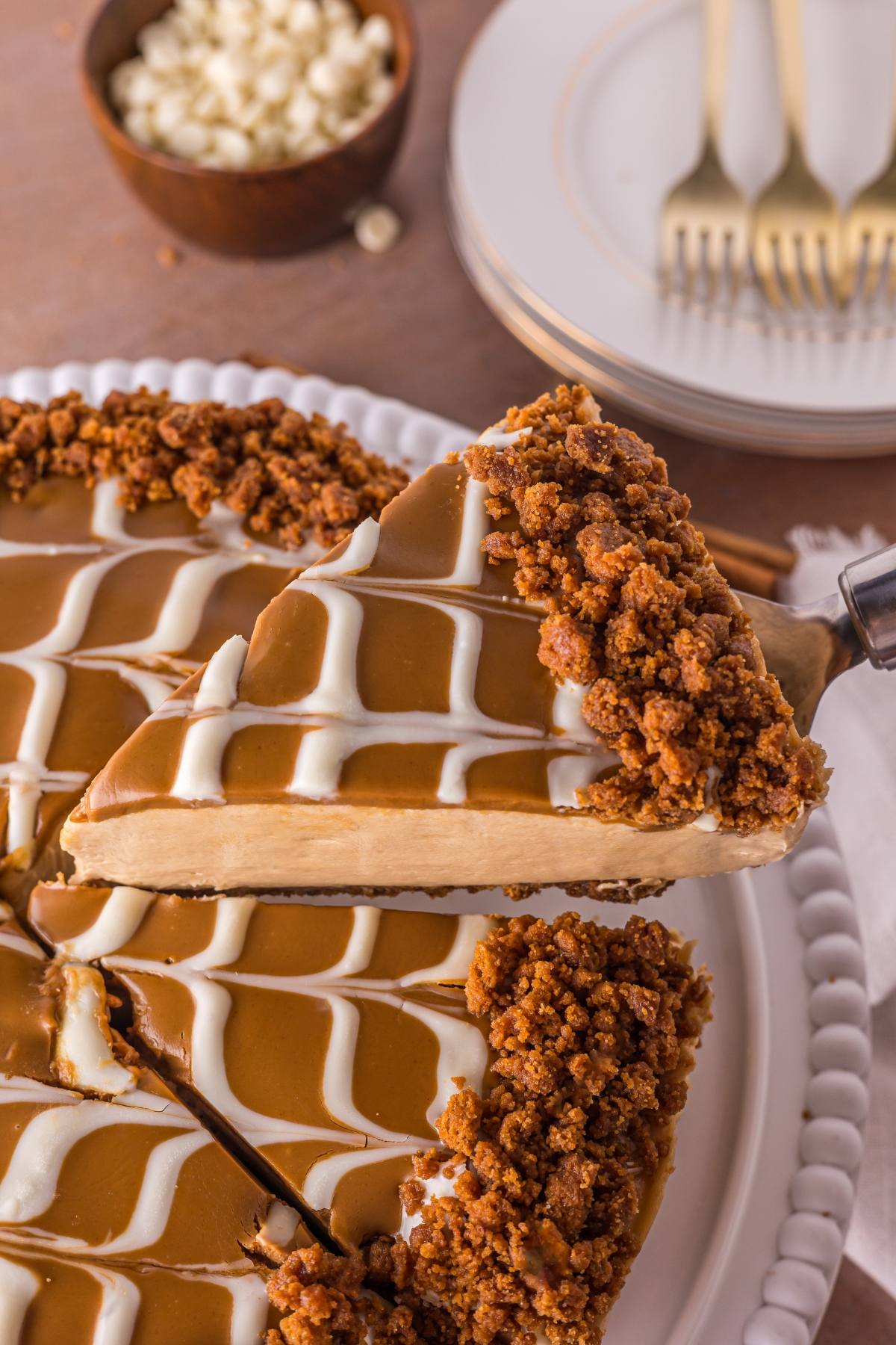 A slice of caramel no bake biscoff  cheesecake with white icing and crumb topping is being lifted from a whole cake.